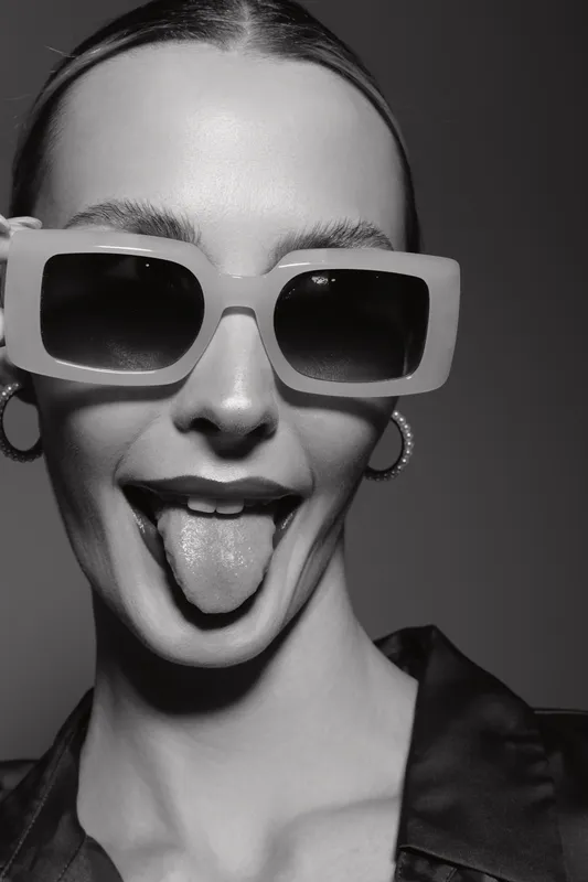 Young white woman with light skin wears large sunglasses and shows tongue in playful black and white portrait.
