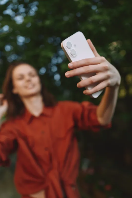 Smiling woman in a red shirt taking a selfie outdoors in a park. Casual lifestyle moment focused on self portrait, outdoor enjoyment, and relaxed mood with soft natural light