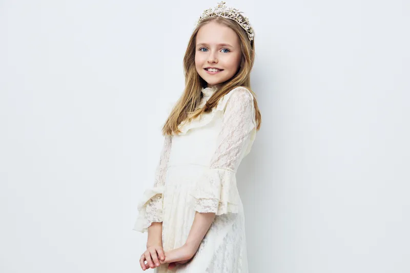 Young girl in a white dress and tiara smiling charmingly, showcasing innocence and joy against a light background, perfect for themed celebrations or events