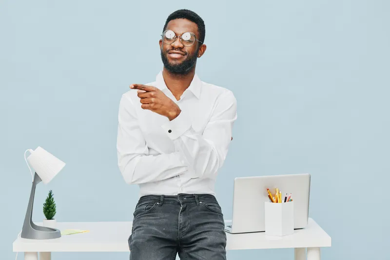 Confident young African American man wearing glasses, white shirt, standing at modern workspace with laptop, pointing sideways, smiling, and looking away against light blue background. Business and co