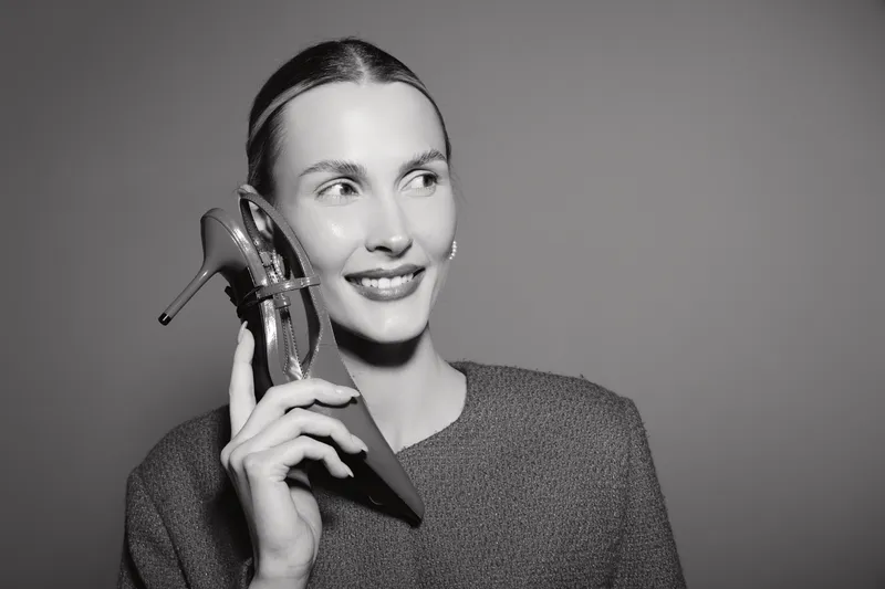 Smiling white woman with light skin holds high heel shoe near face in studio. Concept of fashion and happiness.