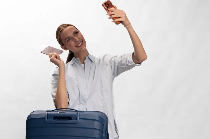 Smiling woman with long hair holding passport and smartphone takes selfie near blue suitcase. Concept of travel, vacation, tourism, and modern lifestyle with happy mood and casual outfit.
