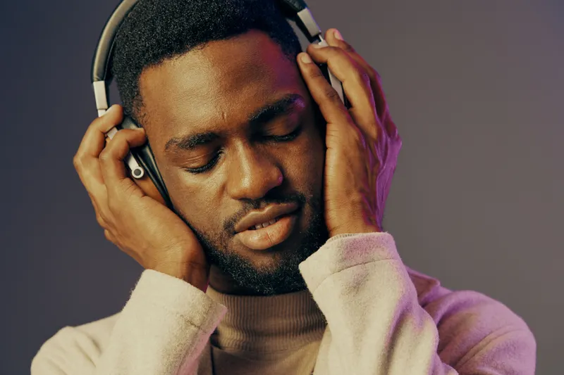 A young African man enjoying music with headphones, showcasing relaxed emotions against a soft gradient background, ideal for themes of relaxation and music appreciation