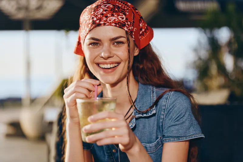smiling woman with red bandana enjoying refreshing drink outdoors in sunny summer setting, casual denim clothing, natural light and warm atmosphere
