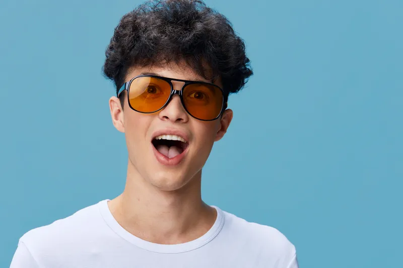Young man wearing orange sunglasses and a white t-shirt, expressing surprise and joy against a blue background in a casual studio portrait.