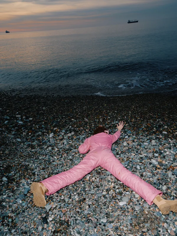 Beach scene with a person in a pink suit lying on pebbles along the shoreline at sunset, capturing stillness and contemplation as calm waves meet the horizon under warm light.