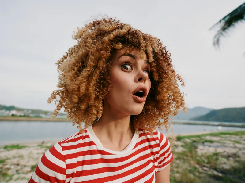 Portrait of a young woman with vibrant curly hair in a red and white striped shirt, surprised expression, standing outdoors by a calm lake and distant hills.