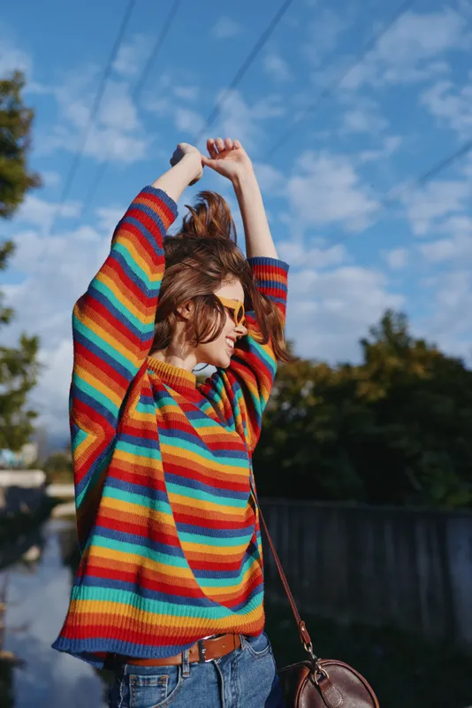 Woman in a colorful striped sweater and sunglasses stretches arms outdoors under a blue sky, smiling in casual outfit with leather shoulder bag for travel and lifestyle.