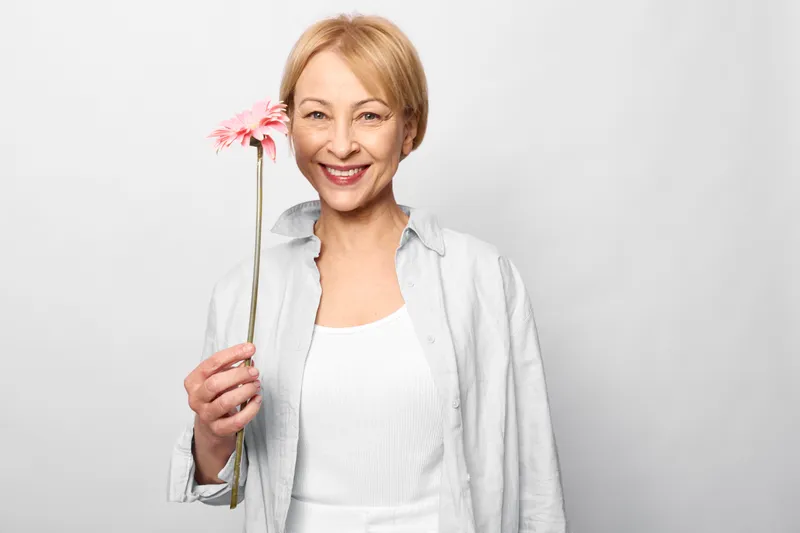 Smiling senior woman holding flowers bouquet in a studio portrait, white background with soft light. Joyful spring mood, freshness and wellness, relaxation, self care concept.