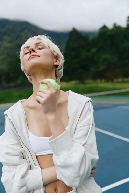Young athletic woman enjoying a healthy lifestyle, holding a green apple outdoors, wearing a light gray hoodie, exuding confidence and joy on a tennis court