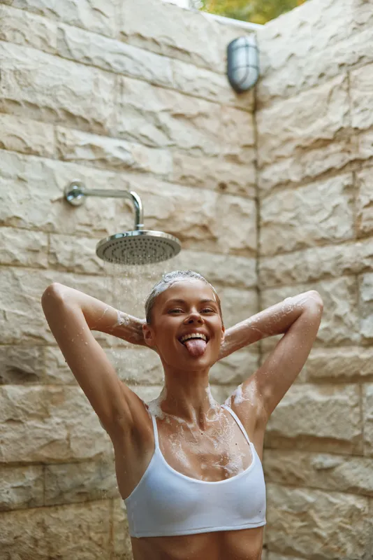 playful woman showering outdoors under rain showerhead with stone wall background, enjoying refreshing water on a bright sunny day