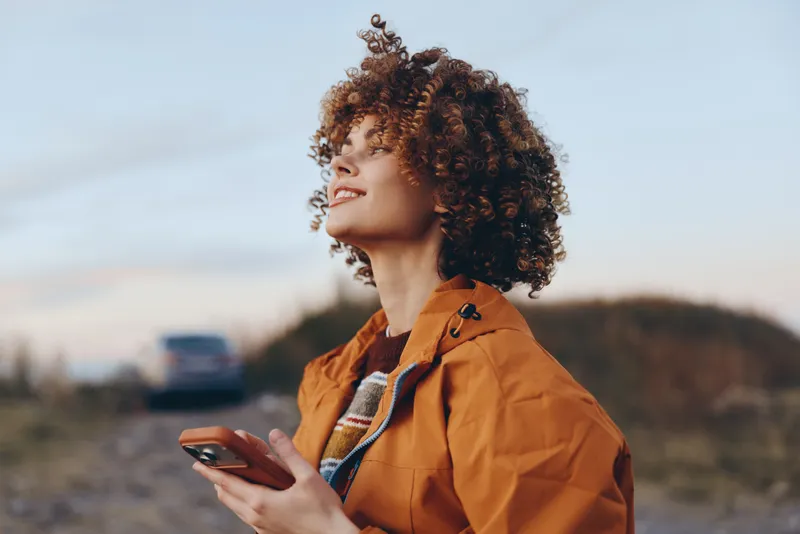 Woman lifestyle portrait outdoors, curly hair and orange jacket, holding smartphone while looking sideways, communication moment in fresh air, soft sunlight and blurred hills softly