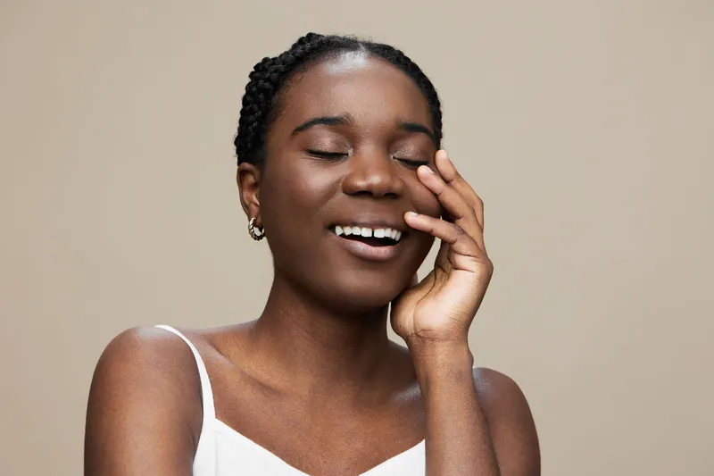 Joyful young Black woman smiling with eyes closed, wearing a simple white top, against a soft neutral background, radiating happiness and confidence