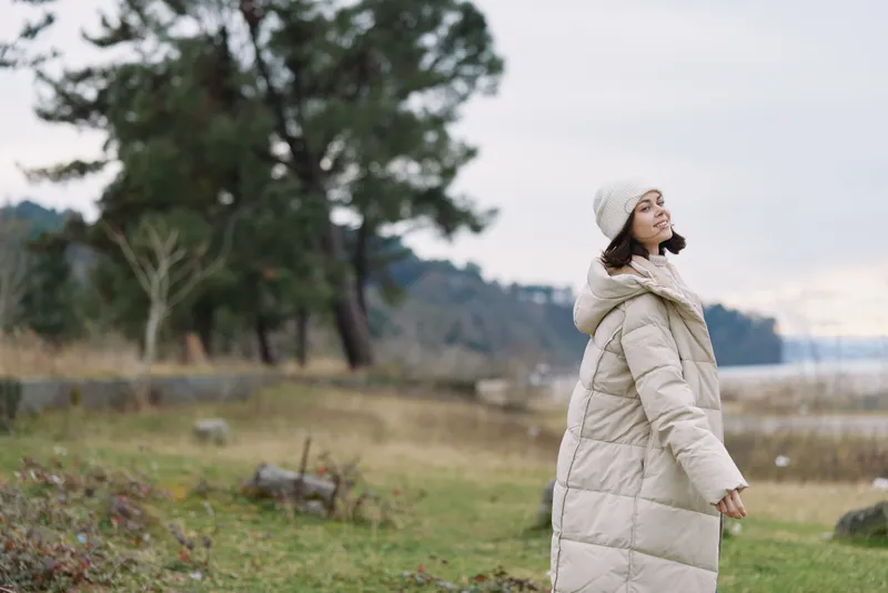 Woman in a long beige coat and knit hat outdoors by a lakeside, trees in the background, capturing a peaceful seasonal moment in nature, calm breeze