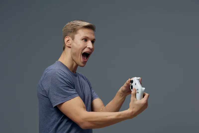 Young Caucasian man with short blond hair playing video game, holding game controller with excited and surprised expression, wearing casual blue t-shirt, standing against plain gray background, studio