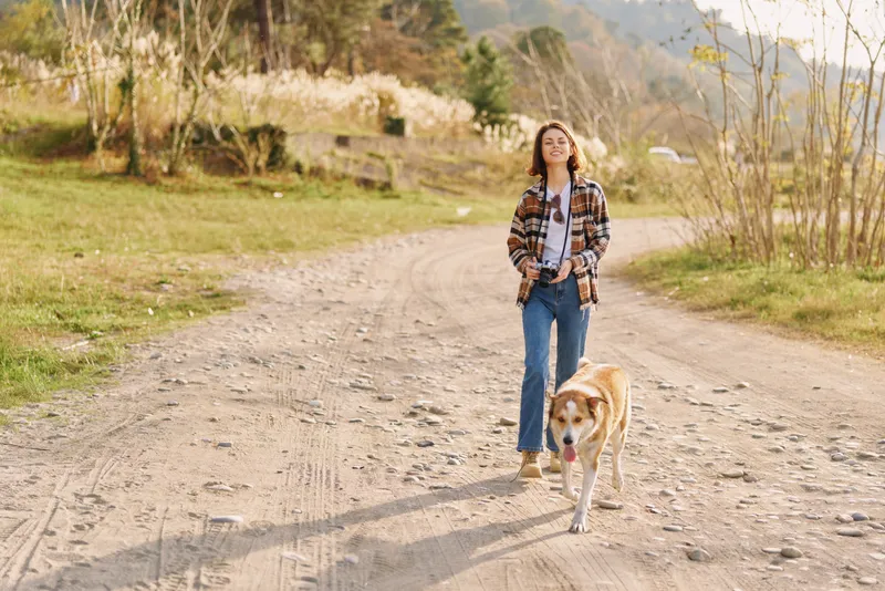 Woman walking a dog along a dirt path in a sunny countryside setting, casual attire, trees and open space, outdoor exercise and companionship in a peaceful nature scene.
