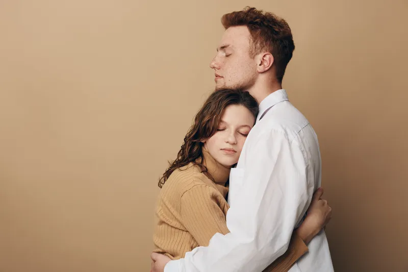 Young couple embraces tenderly with eyes closed against beige background. The woman wears a brown sweater and the man a white shirt. Concept of love, comfort, and emotional connection.