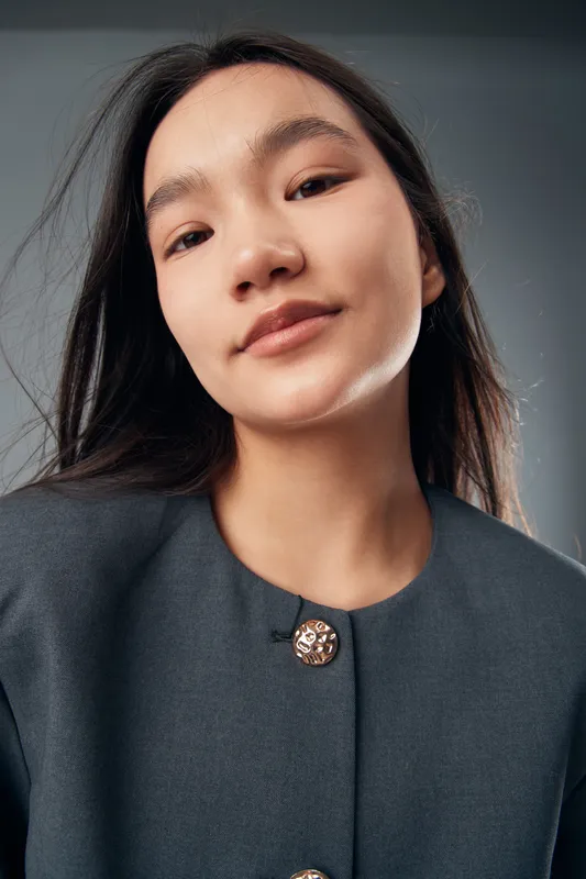 Young woman with long dark hair and natural makeup wearing dark buttoned jacket looking confidently at camera with neutral background and soft lighting in portrait closeup.