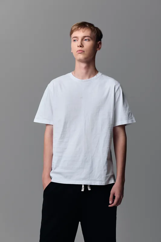 Man wearing a white tee stands in a studio for casual fashion and lifestyle shoot, relaxed pose, gray backdrop, clean and minimalist look suitable for everyday wear.
