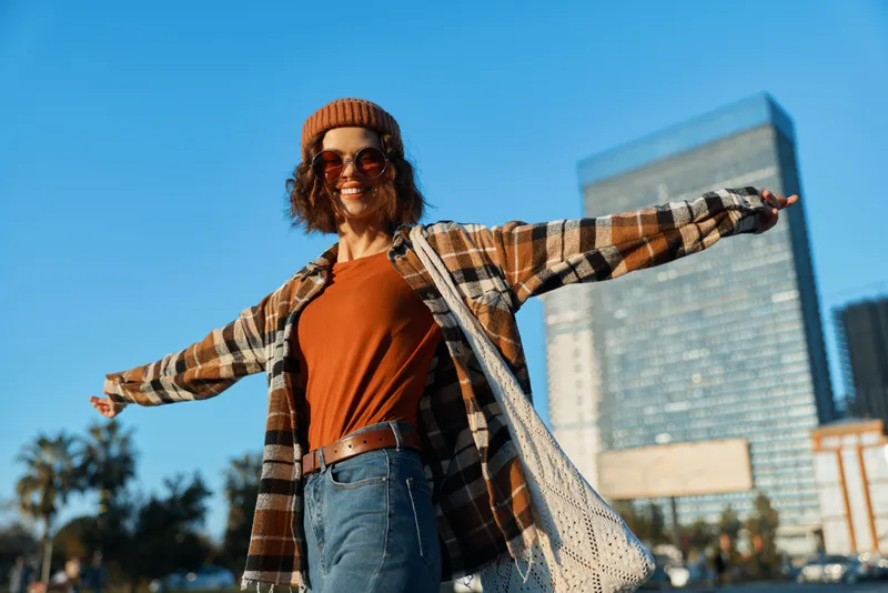 Woman smile in city outdoors wearing beanie and sunglasses, casual outfit with arms outstretched, joy and sunlight, authenticity and candid lifestyle with golden hour glow and emotional storytelling.