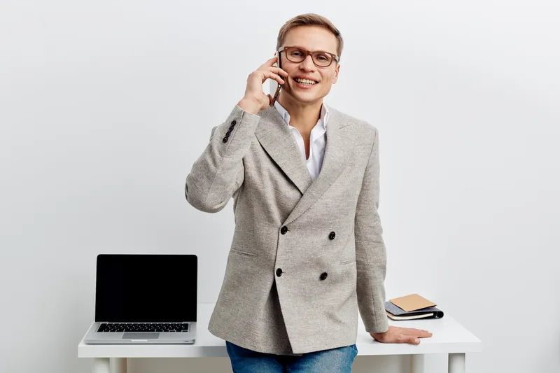 Confident professional woman with glasses talking on mobile phone while standing at desk with laptop and notepad, smiling, office background. Business lifestyle concept.