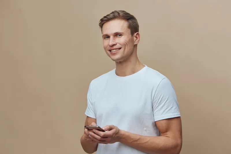 smiling man using mobile phone in white t-shirt look camera hold smartphone on beige background digital nomad concept