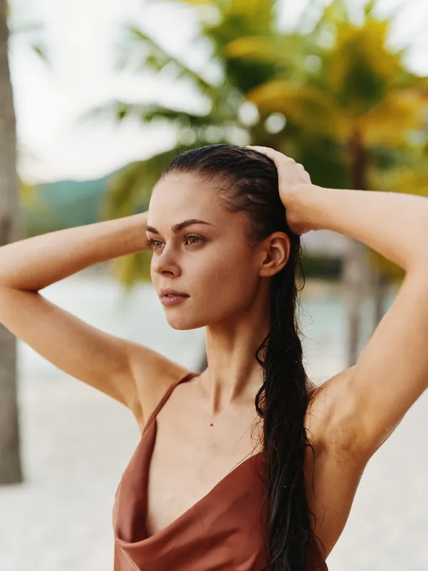Portrait of a young woman on a sunlit beach, hands lifting hair, wearing a brown satin swimsuit top, thoughtful gaze, warm skin tones, palm trees in soft background.