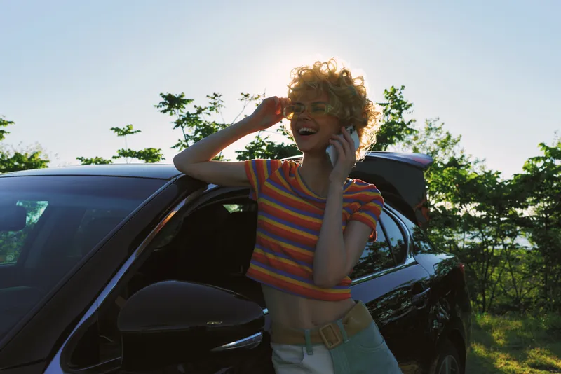 A smiling woman stands beside a car in bright outdoor light, wearing a striped top. A retro, sunny lifestyle moment captures freedom and casual joy, with vibrant color tones.