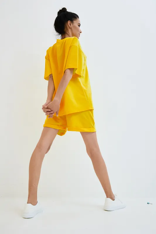 Woman in bright yellow casual outfit stands with hands behind back, wearing white sneakers, minimalist studio background, modern fashion style, relaxed pose.