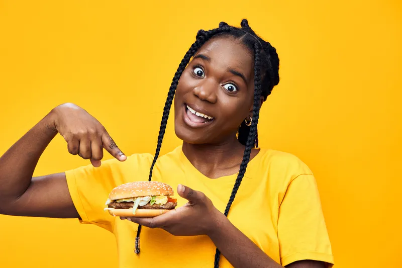 Happy woman enjoying a delicious burger against a vibrant yellow background, showcasing her excitement for tasty fast food with joyful expression