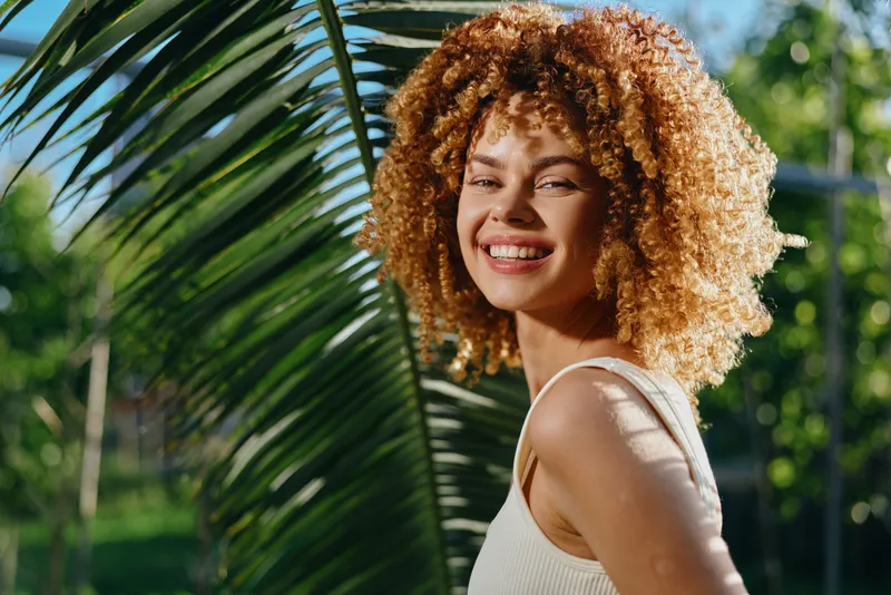smiling woman with curly hair enjoying sunny tropical outdoors, natural light, vibrant green palm leaves background, summer mood, casual white top