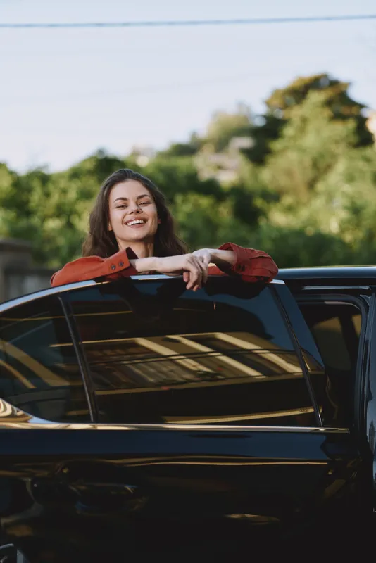 A young adult woman leans from a glossy black car window with a bright smile, embodying a carefree street style moment in the sun, the candid pose and open mood convey spontaneity, confidence