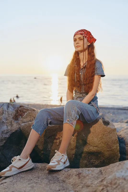 young woman sitting on rock at sunset by the sea wearing casual outfit and red bandana, relaxed mood with natural light and summer atmosphere