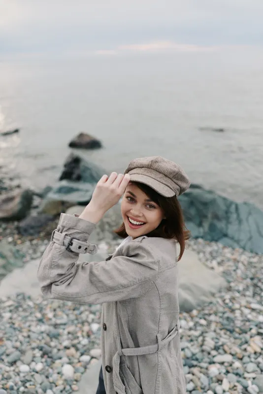 Smiling young woman in stylish coat and hat poses on rocky beach near calm sea. Natural light highlights happy expression and autumn fashion. Concept of outdoor lifestyle and travel.