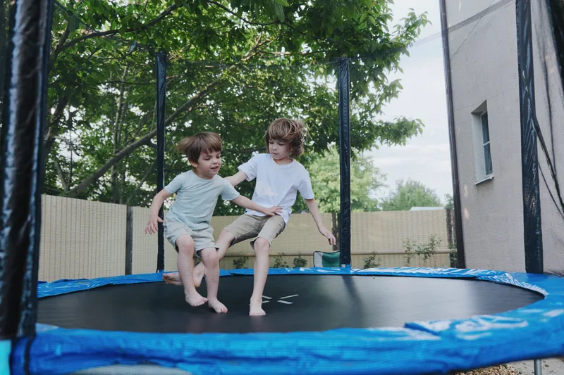Two happy boys jumping on a trampoline in a sunny backyard, showcasing fun and playfulness in a vibrant outdoor setting, surrounded by green trees.