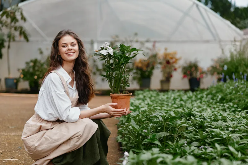 Happy young woman in a greenhouse holding a potted flower, wearing casual clothing, surrounded by lush greenery, representing a vibrant gardening concept