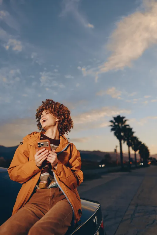 Woman with curly hair holding smartphone on street at sunset, outdoors lifestyle, orange jacket, palm trees and mountains in background, warm evening light, casual travel portrait