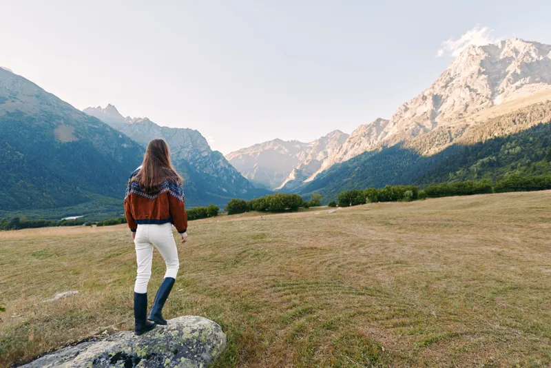 Woman hiking in mountains, landscape and meadow, wearing sweater and boots, standing on rock and looking at distant peaks. Travel, adventure and nature exploration in scenic outdoors.