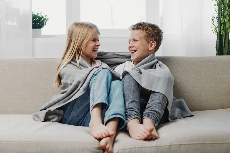 Two happy children wrapped in a gray blanket sit on a beige sofa, smiling and looking at each other in a bright living room. Concept of warmth, friendship, and cozy home moments.