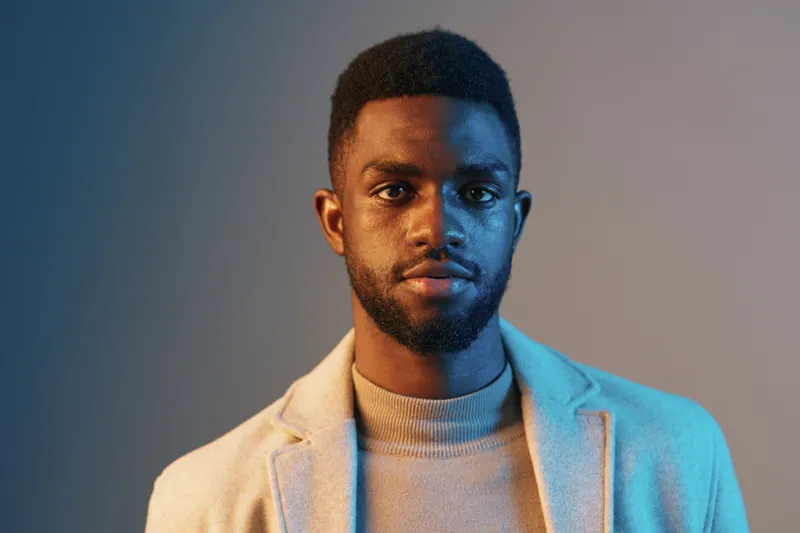 Stylish African American man in a light blazer and turtleneck with a confident expression against a gradient background, showcasing modern fashion and sophistication