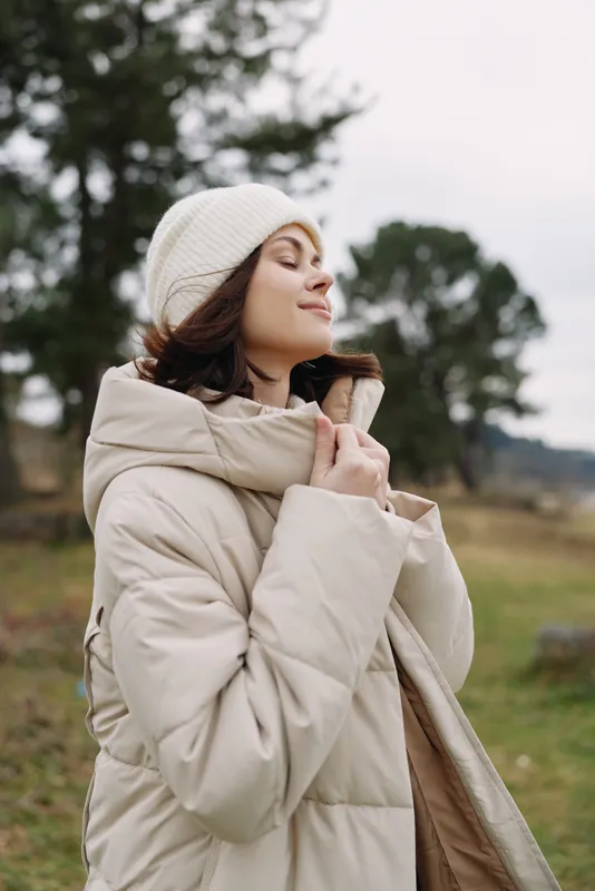 Woman in a beige puffer coat and knit beanie enjoys a chilly outdoor moment with a calm smile amid open nature
