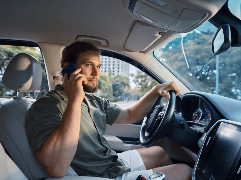 Portrait of a man driving a car while talking on a mobile phone, wearing a casual green shirt, focused expression, hands on the wheel, dashboard and rearview mirror visible.