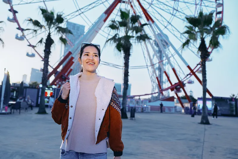 Smiling woman wearing a warm jacket at an outdoor amusement area near a large ferris wheel and palm trees during twilight, capturing a cheerful moment and a carefree travel atmosphere.