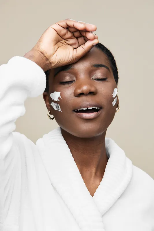 Beautiful African woman applying skincare cream, smiling with eyes closed, wearing a white robe, showcasing a fresh and radiant complexion, perfect for beauty concepts