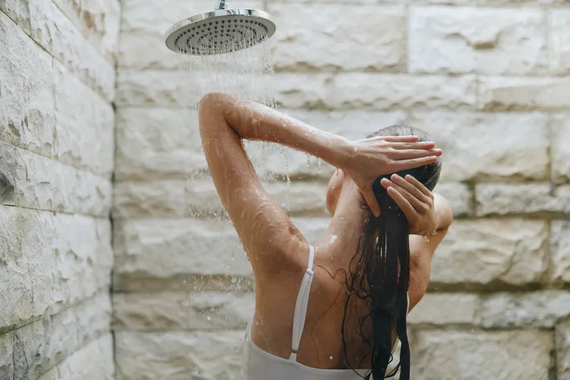 woman showering under rain shower with wet hair standing in modern stone wall bathroom, natural cleansing moment, wellness and relaxation concept
