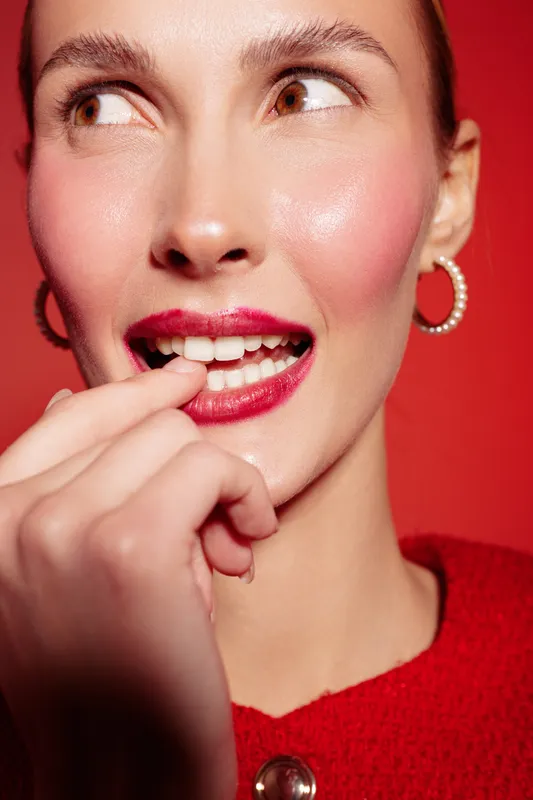 Close-up of a woman with red lipstick and hoop earrings biting her finger, looking sideways. Bright red background and sweater create a bold, stylish portrait concept.