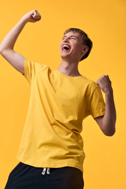 Energy, celebration, joy, enthusiasm, victory, youth illustrated by a smiling male in a yellow T shirt raising his fists in triumph against a bright backdrop, conveying upbeat mood and motivation for