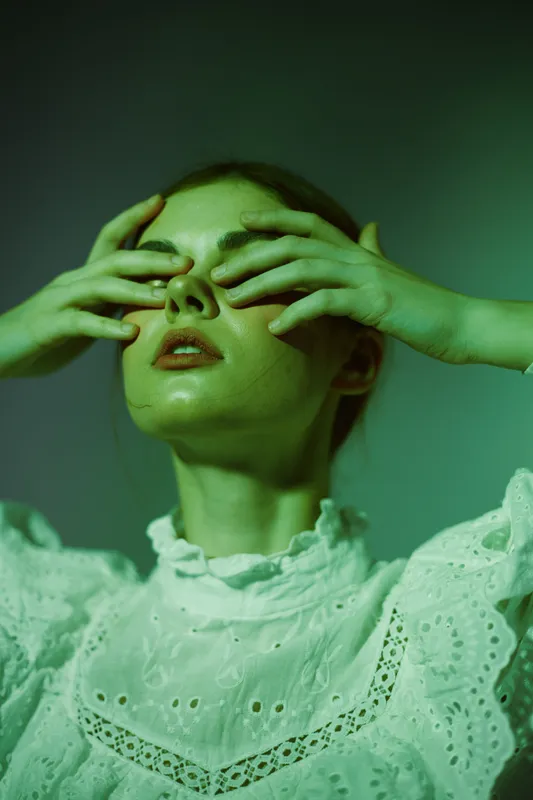 Young woman with a serene expression covering her eyes, wearing a vintage white dress, set against a green background, evoking feelings of tranquility and introspection
