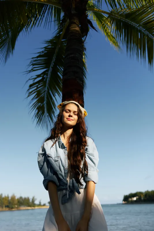 Smiling woman standing under a palm tree near the beach, wearing casual clothes and flowers in hair, enjoying a sunny tropical day by the ocean.