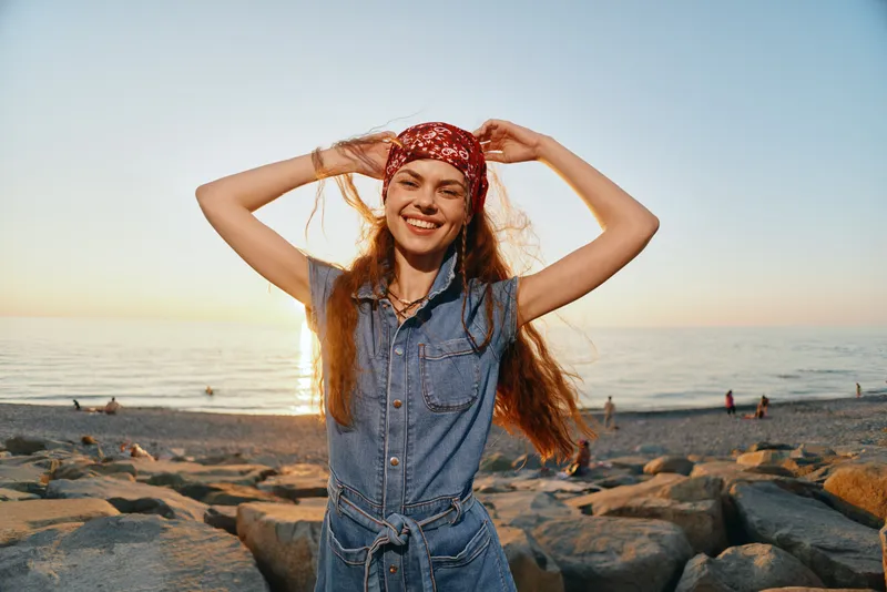lifestyle woman in modern boho-western denim street style smiling happily at the beach with film color sunset background and relaxed vibe